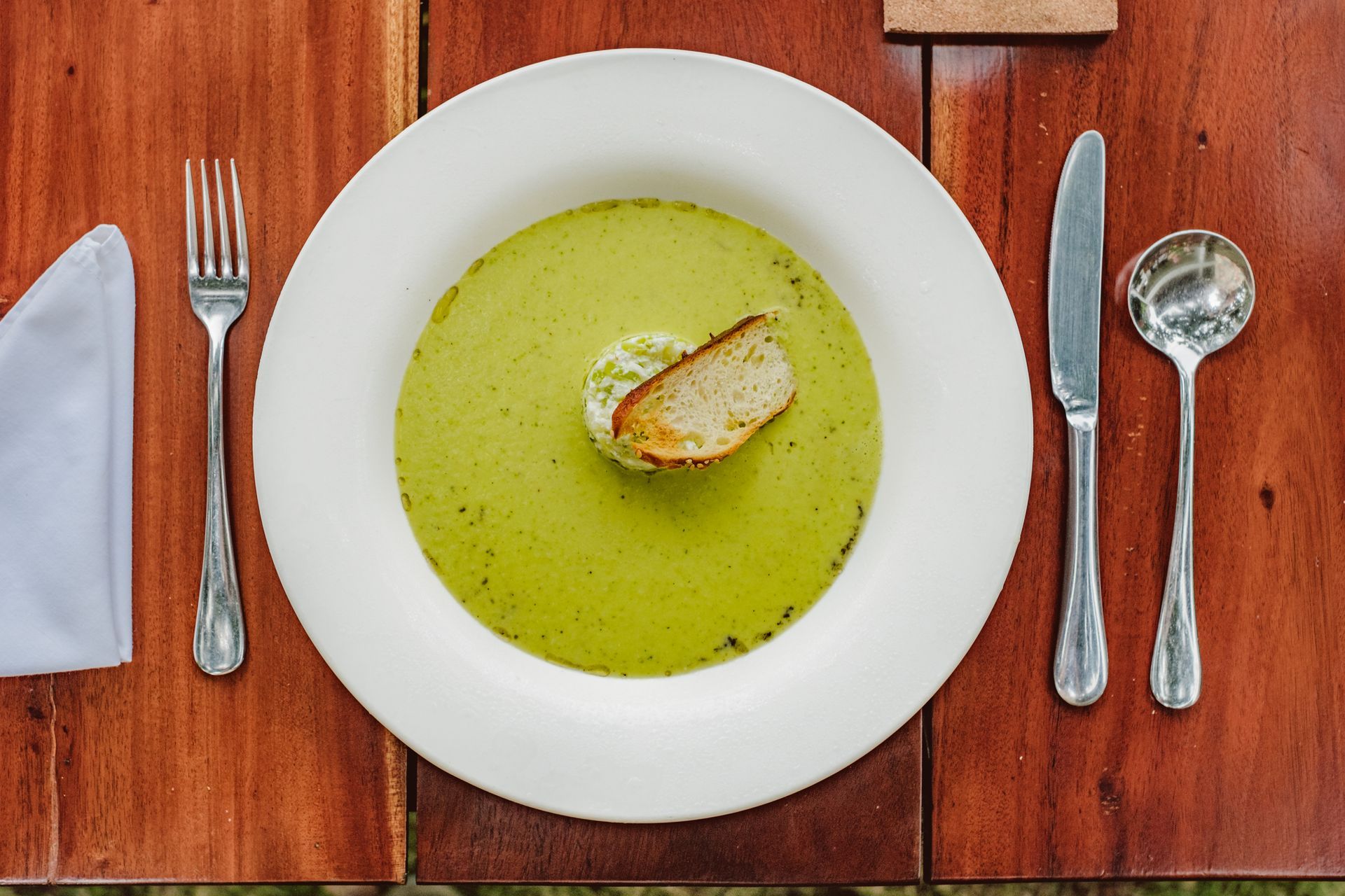 Bowl of green soup with bread on a wooden table, flanked by fork, knife, spoon, and folded napkin