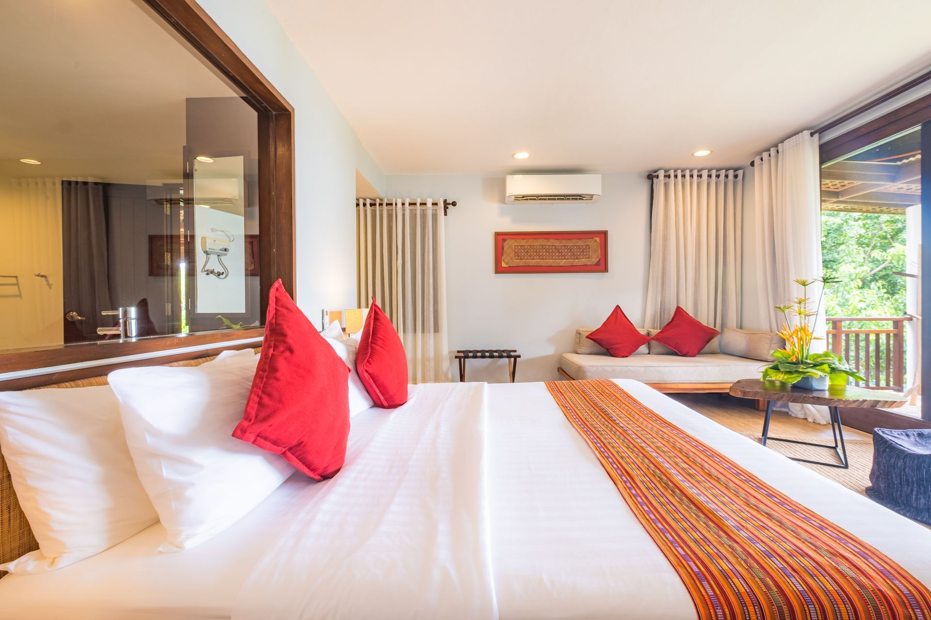 Bright hotel room with king bed, red pillows, wall art, sofa, curtains, and balcony view