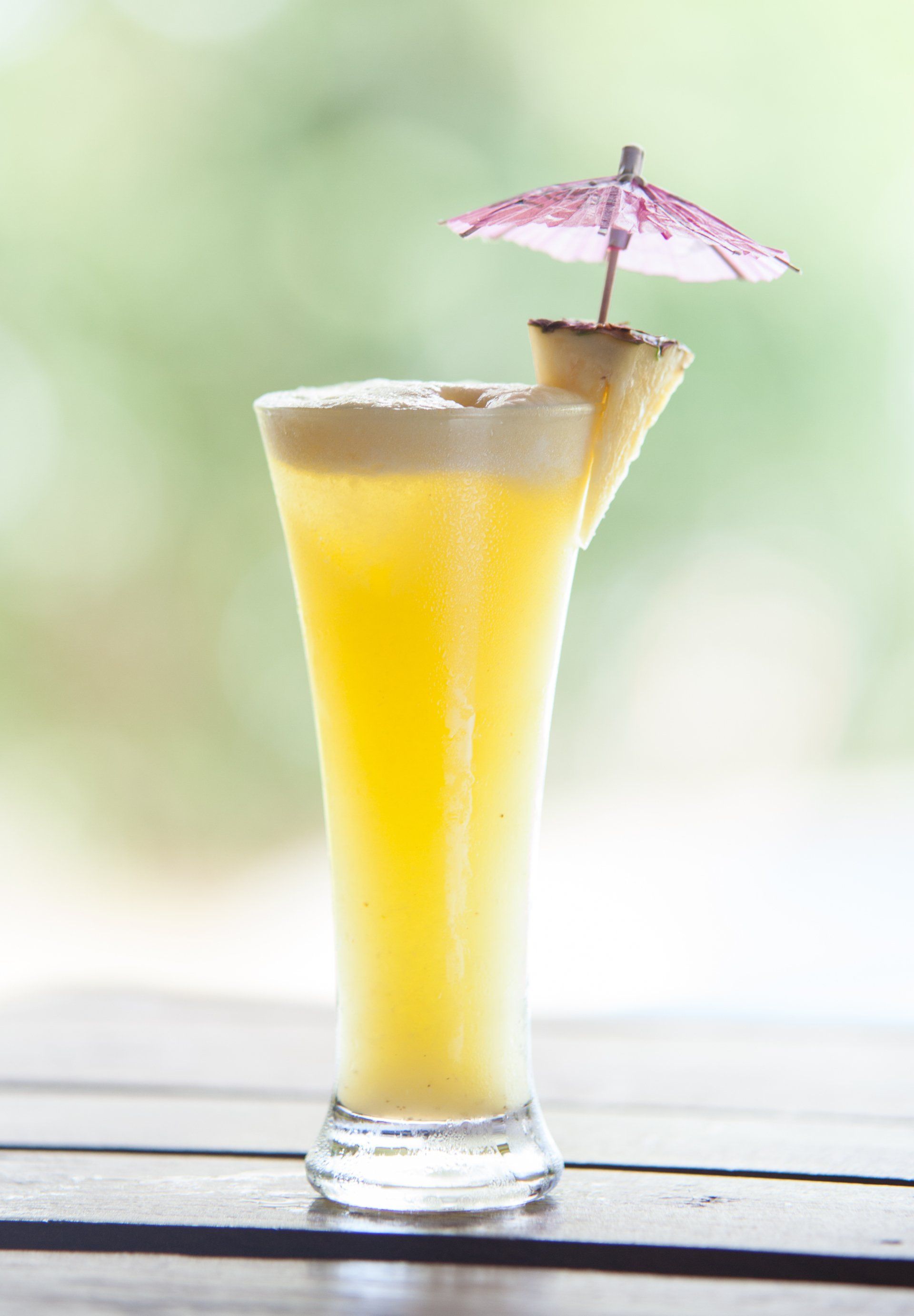 Yellow tropical drink in a tall glass with a pineapple wedge and pink paper umbrella