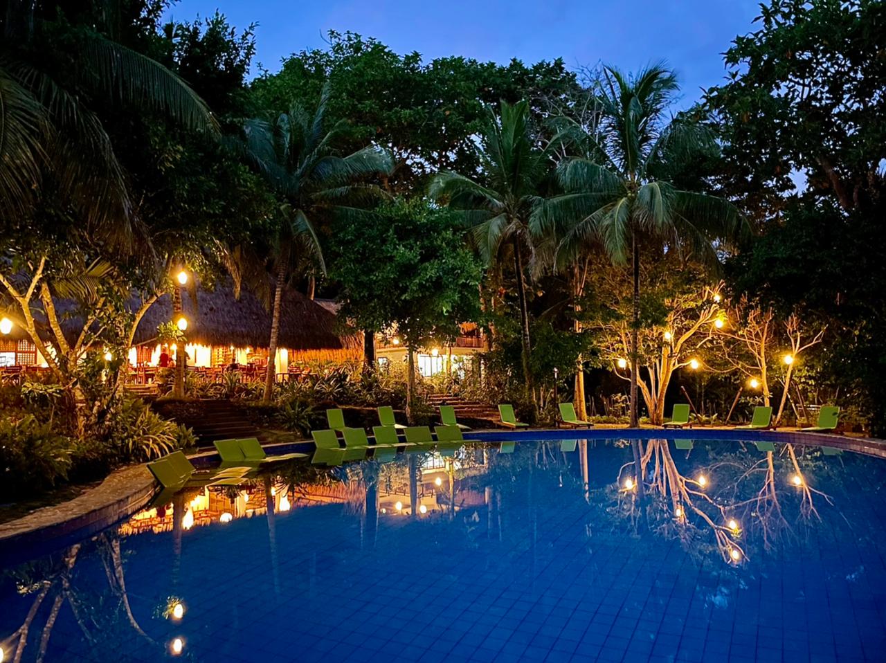 Tropical resort pool at dusk with glowing lights, palm trees, and lounge chairs reflecting in blue water