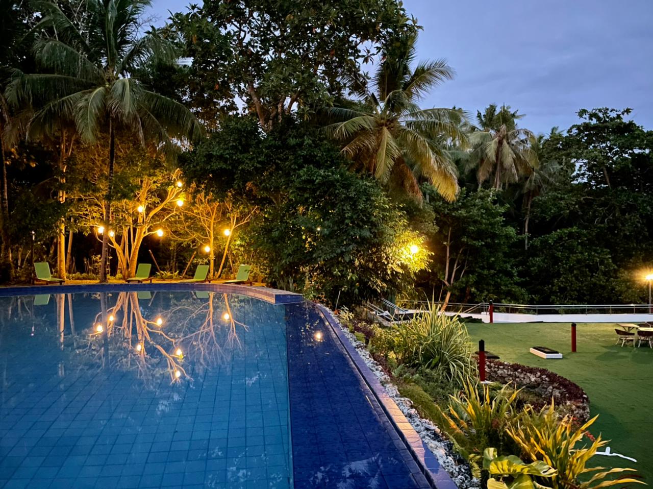 Tropical pool at dusk with blue water, palm trees, warm garden lights, and a lush green lawn.