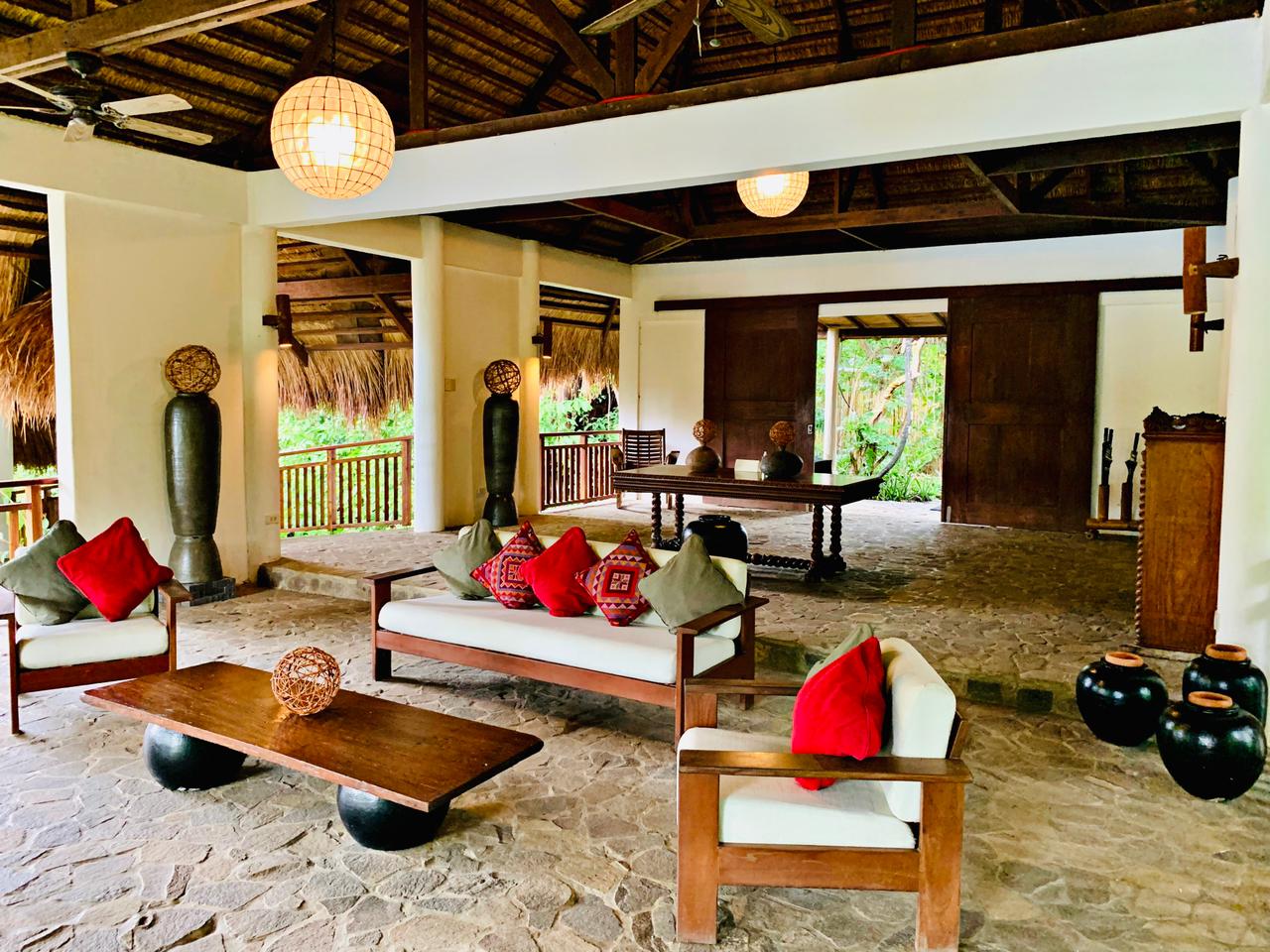 Open-air living room with wooden furniture, red cushions, and thatched roof in a tropical setting