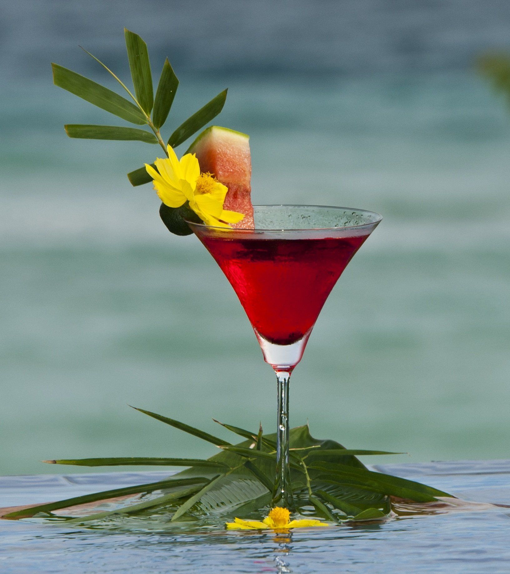 Red martini in a stemmed glass with a pineapple garnish and yellow flower on a beach backdrop