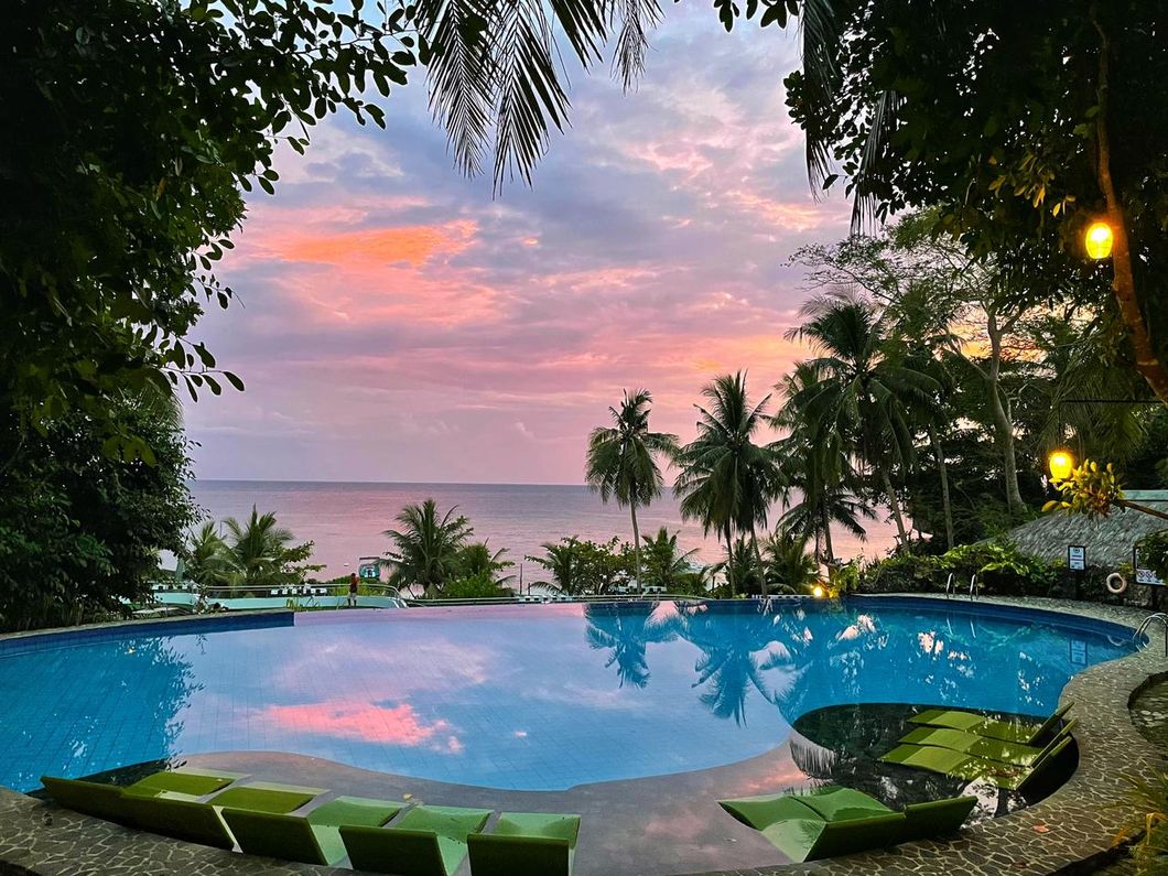 Infinity pool at sunset overlooking the ocean, with palm trees and pink clouds reflected in the water