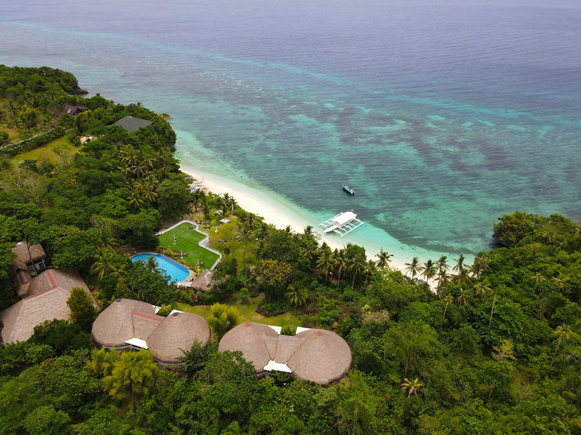 Tropical resort with thatched huts, a blue pool, and a small boat by a turquoise beach