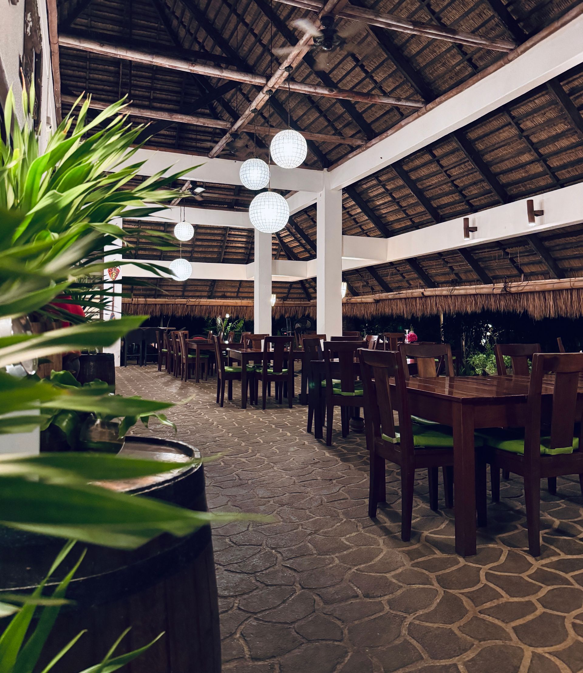 Open-air restaurant with thatched roof, wooden tables, and stone floor, seen through green plants