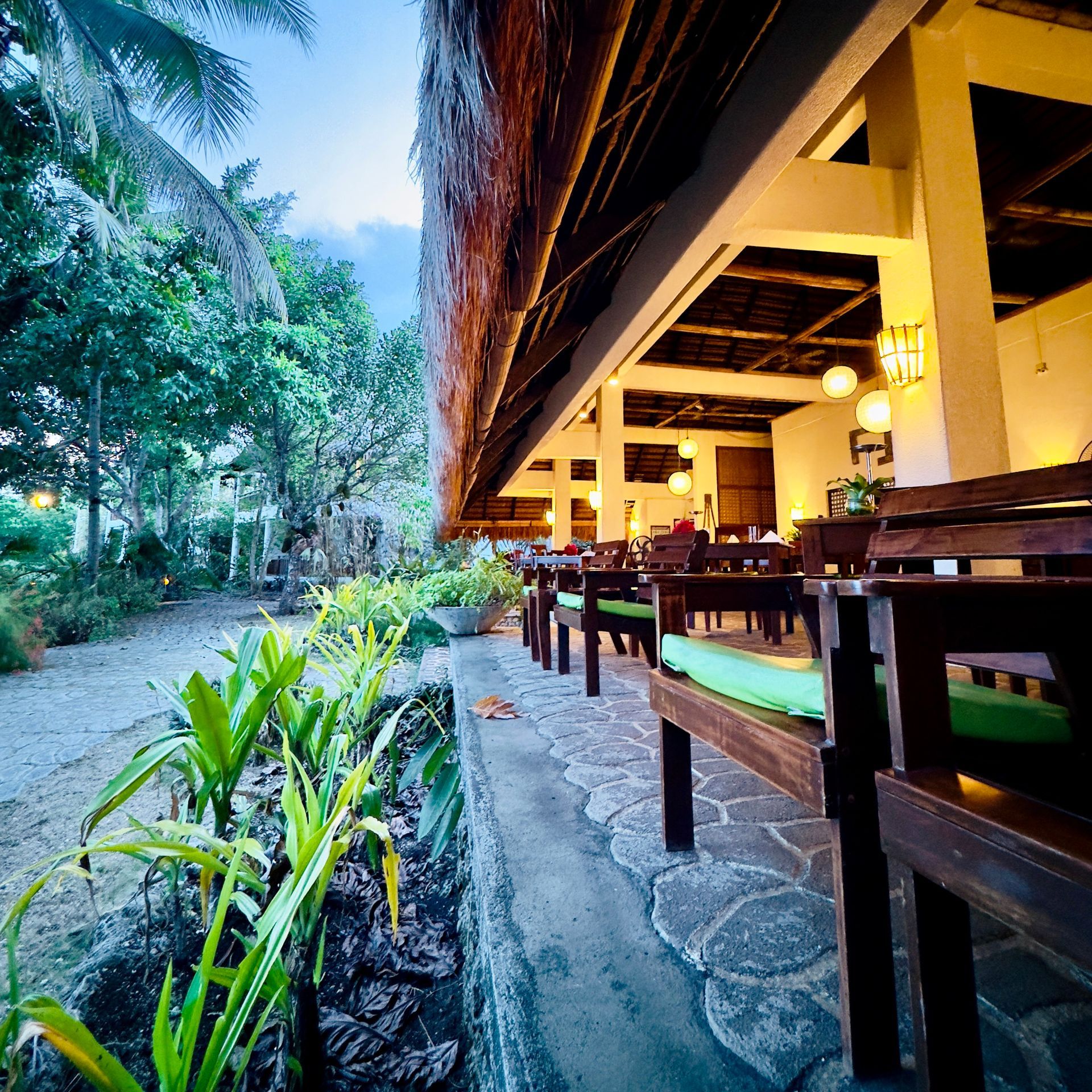 Outdoor restaurant terrace beside a lush garden path, with wooden tables under warm yellow lights at dusk.