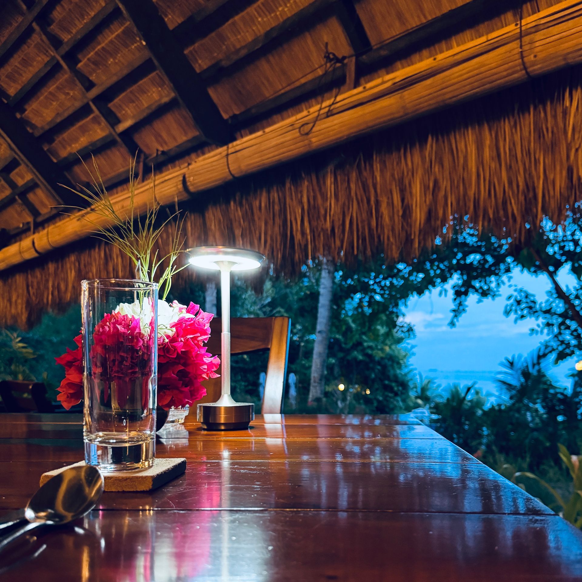 Tropical open-air bar with polished wooden table, pink flowers, and a glowing lamp at dusk