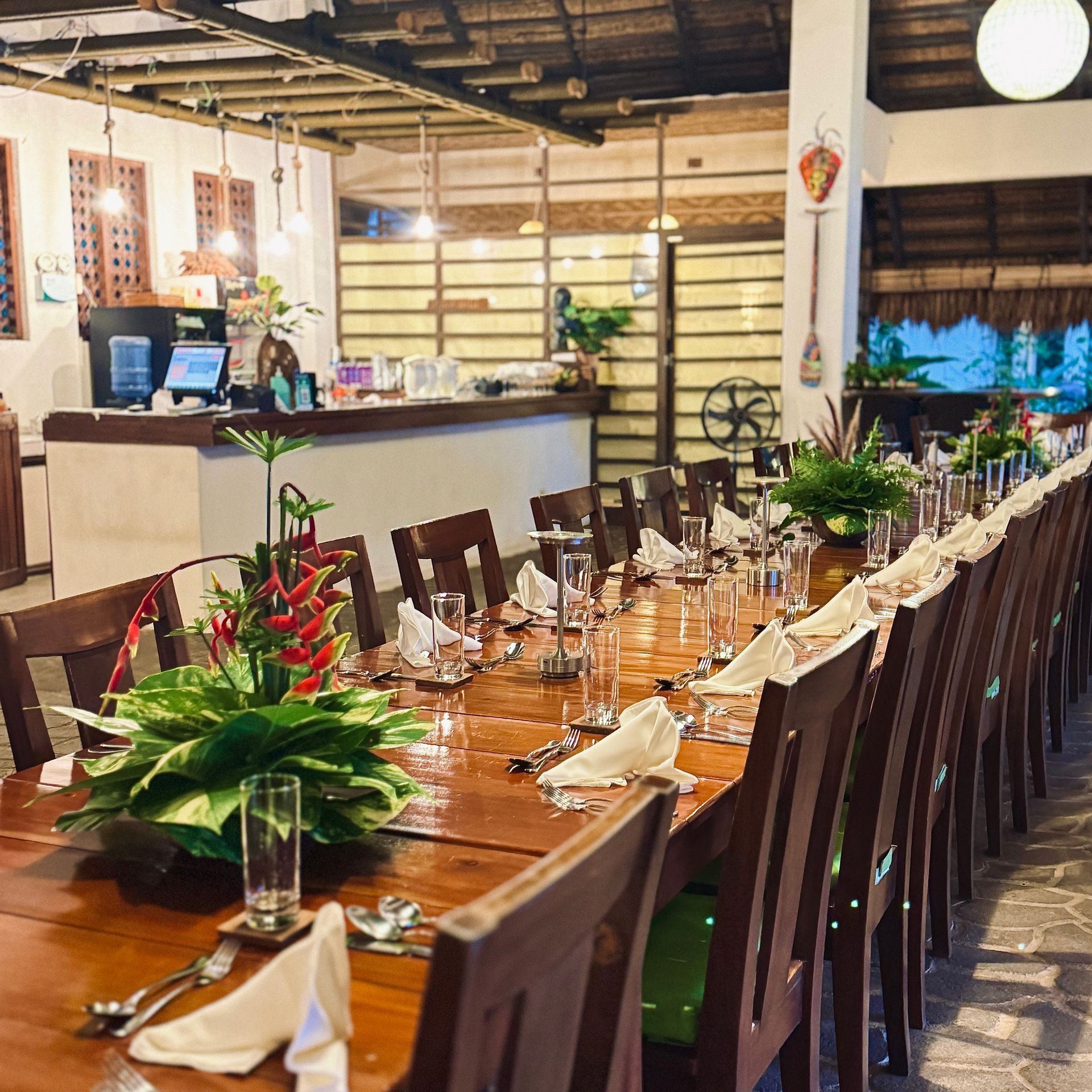 Long dining table set with glasses, plants, and folded napkins in a warmly lit restaurant