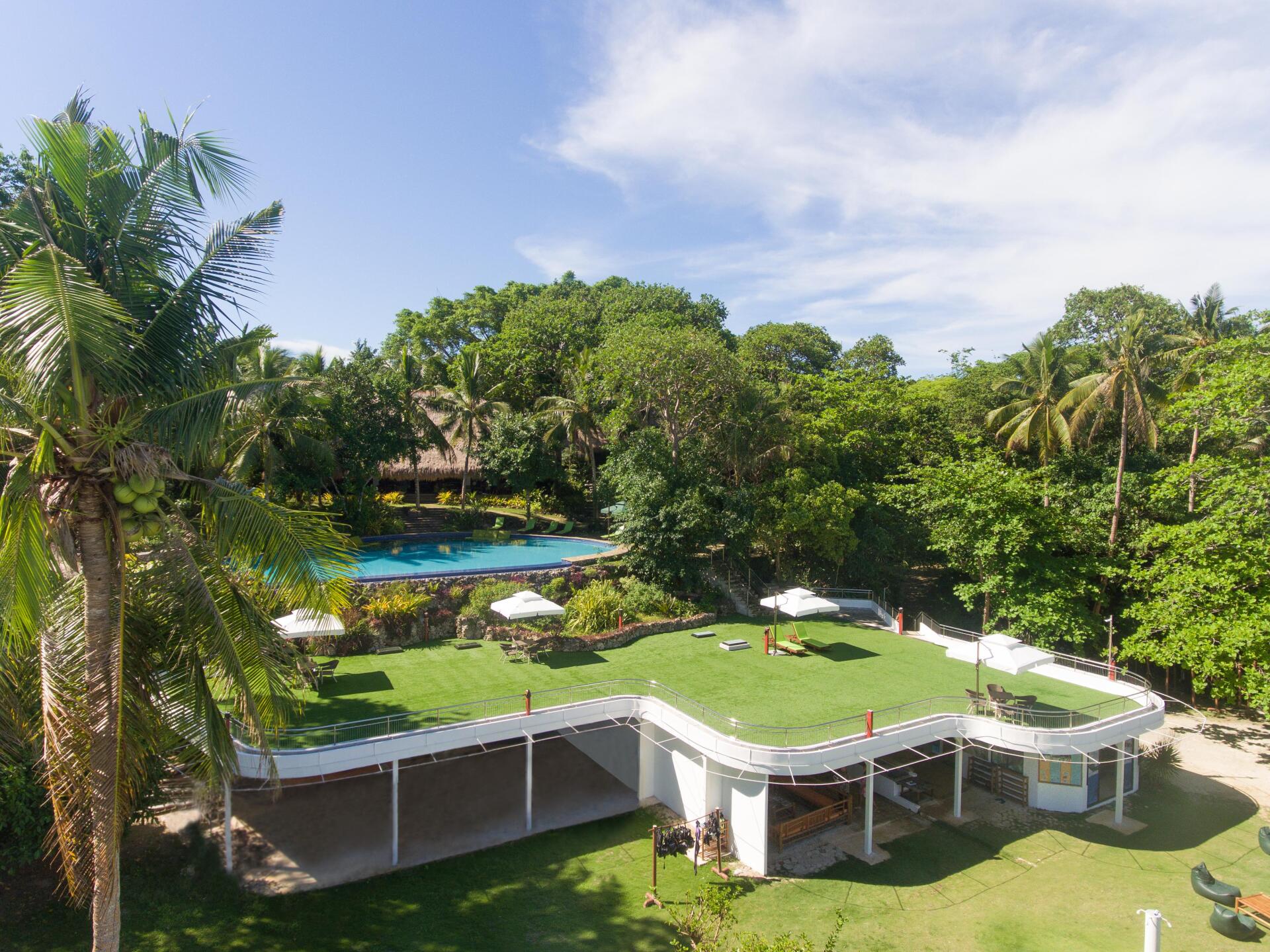Tropical resort with green lawn, white patio umbrellas, blue pool, and palm trees under a sunny sky