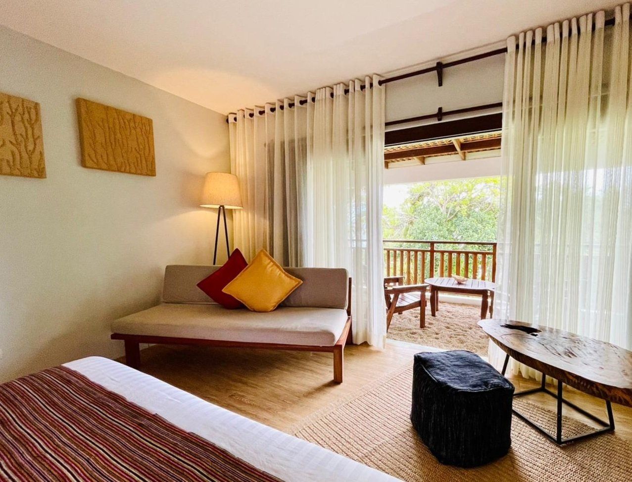 Cozy bedroom with bed, sofa, lamp, curtains, and open balcony view of trees and deck