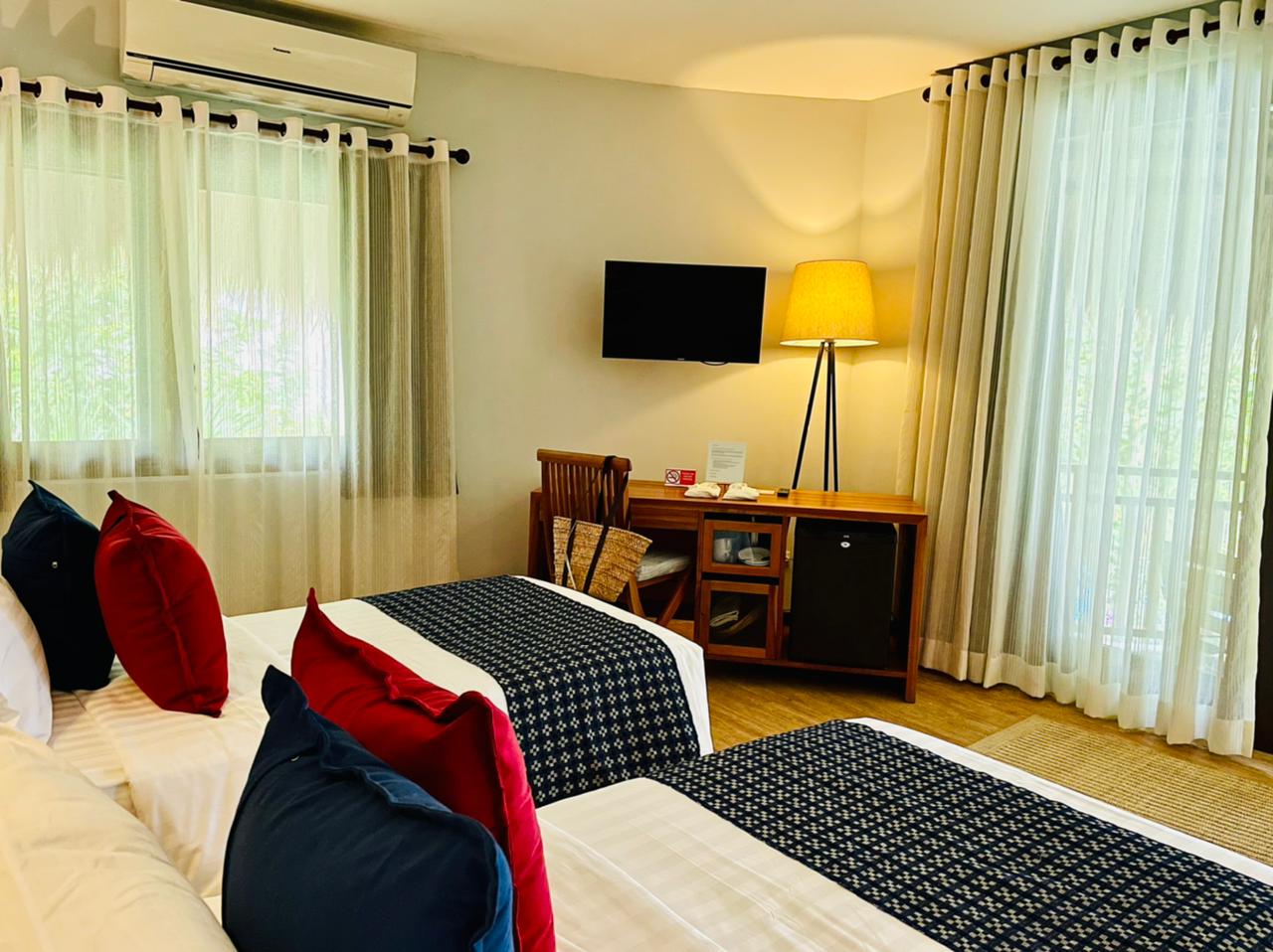 Cozy bedroom with twin beds, wall-mounted TV, desk, and floor lamp by large curtained windows