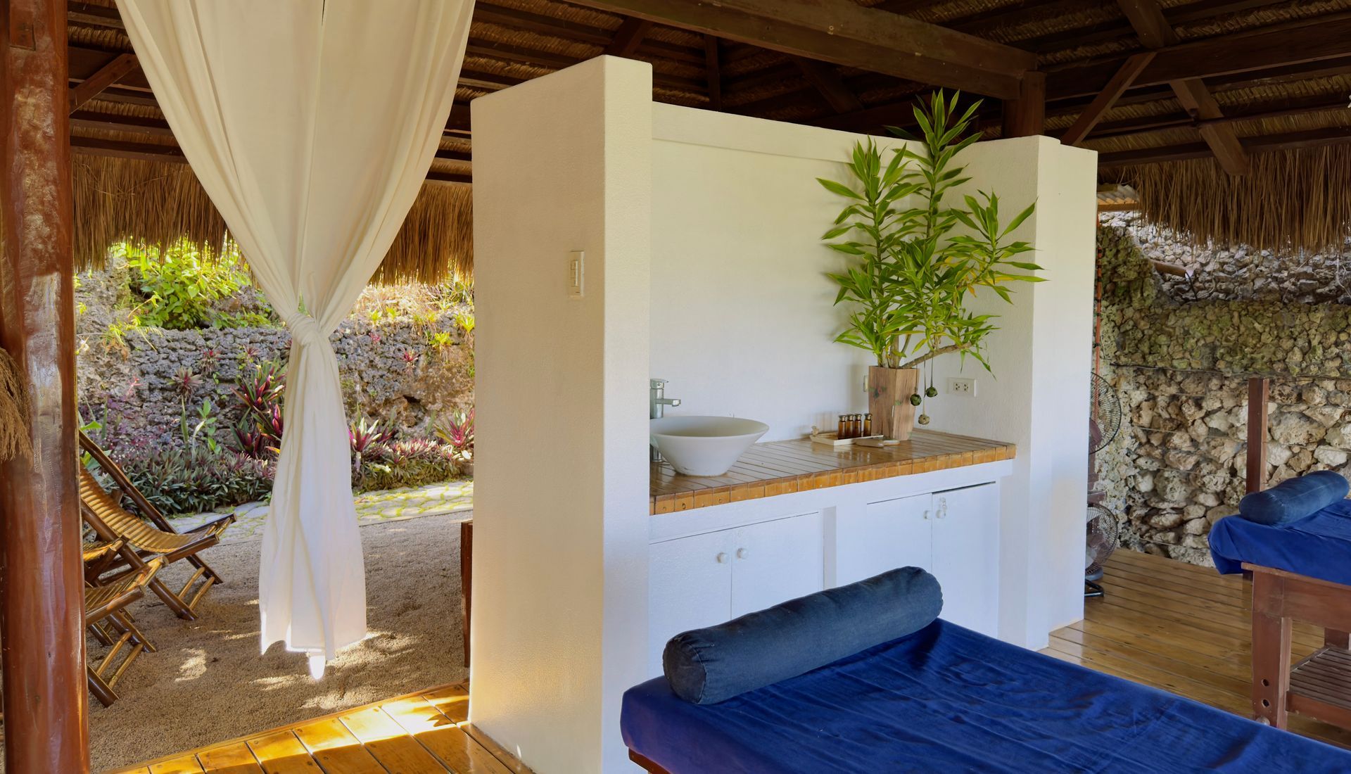 Open-air spa hut with white sink wall, bamboo roof, blue lounge chairs, and curtains.