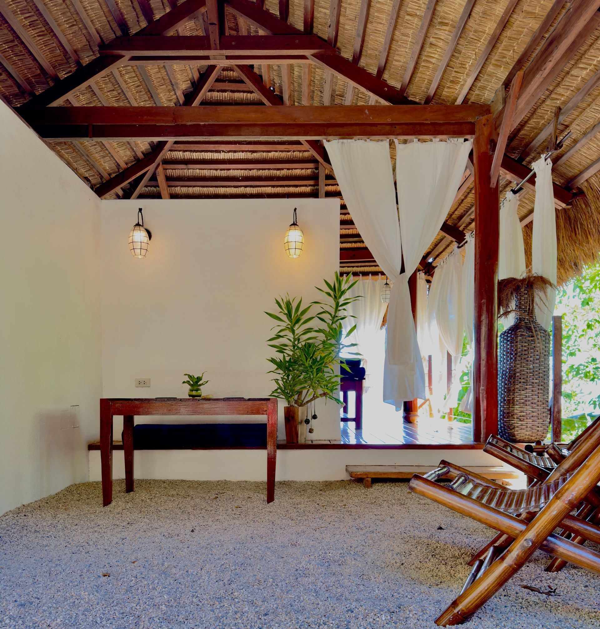 Open-air thatched patio with wooden table, hanging lights, potted plant, and lounge chairs