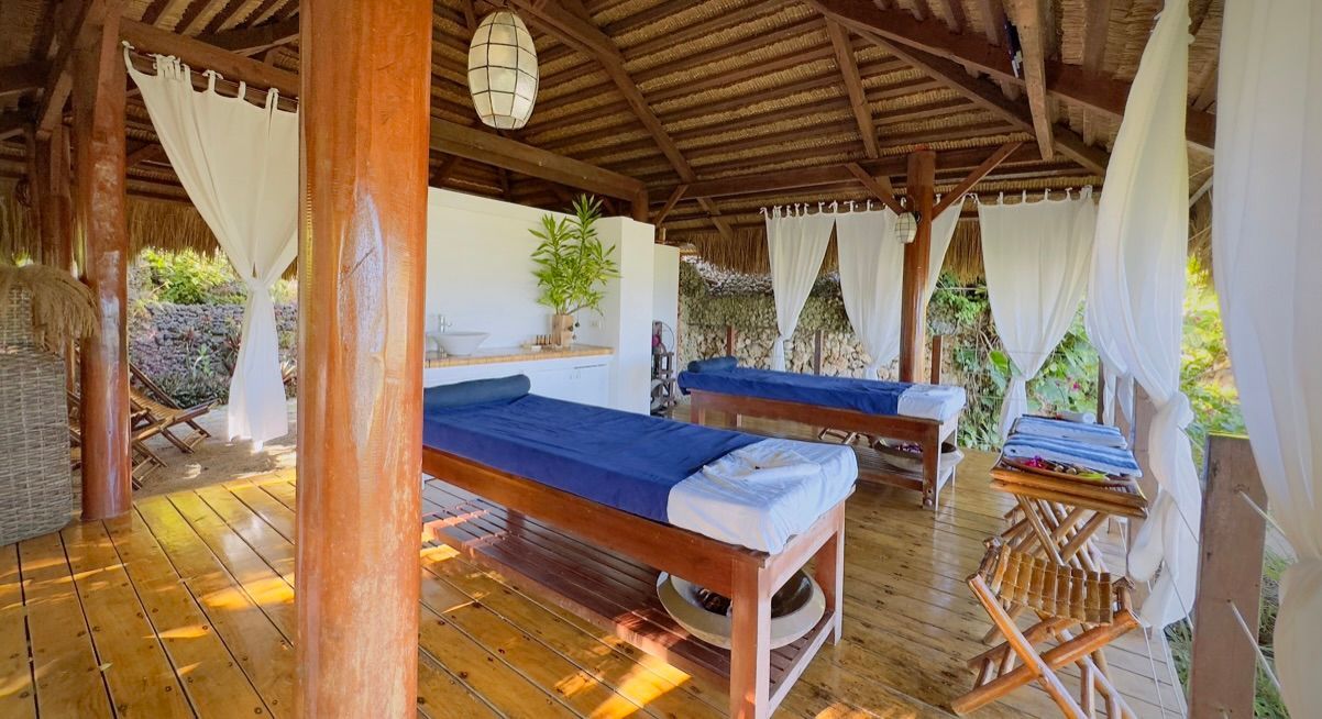 Open-air massage pavilion with two blue-covered tables, wooden posts, and white curtains in a tropical setting