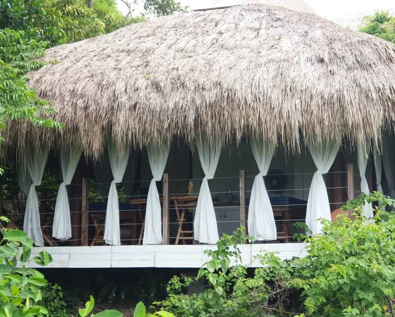Thatched-roof open-air hut with white curtains, surrounded by lush green foliage