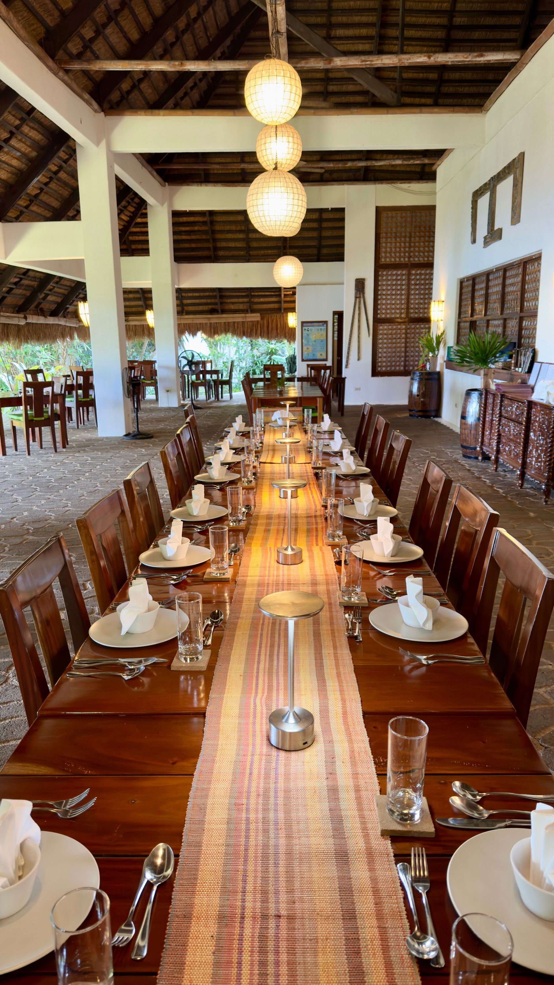 Long wooden dining table set with white plates and glasses in a bright, rustic restaurant hall
