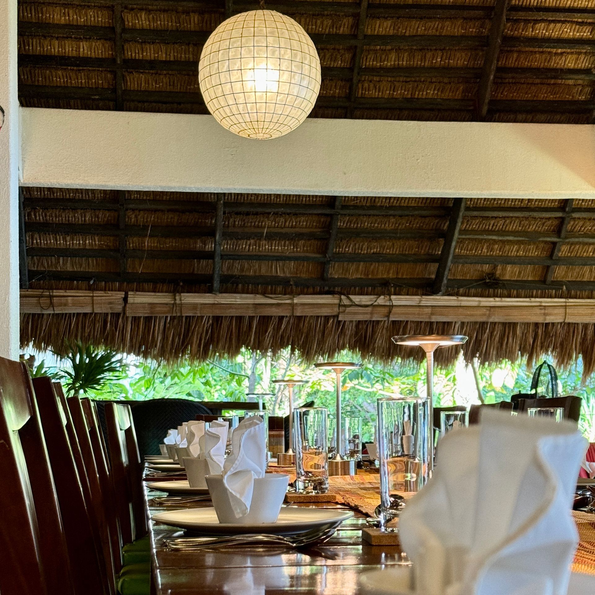 Open-air restaurant table set with white napkins, plates, and glassware under a thatched roof