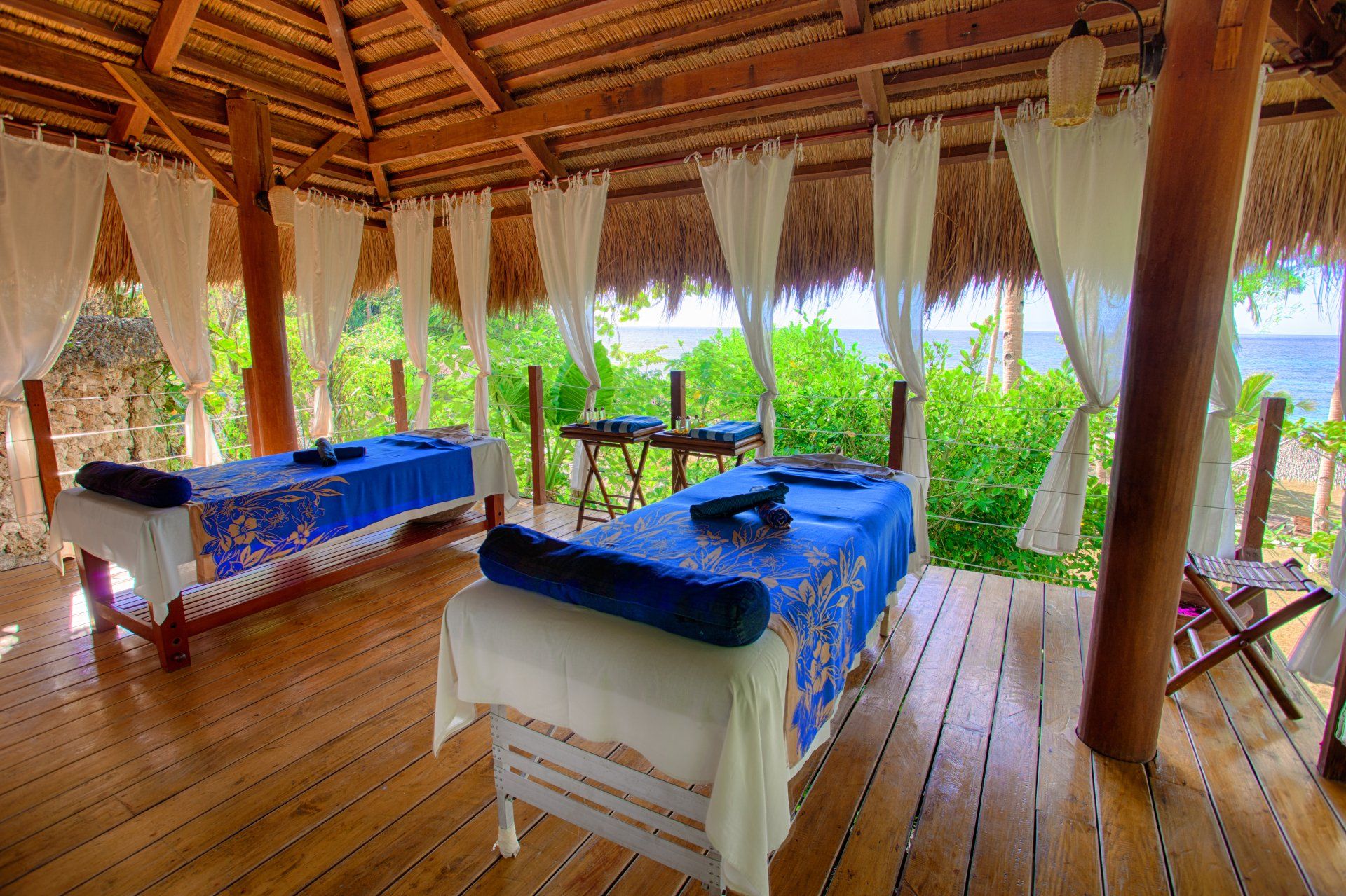 Open-air spa room with two blue massage tables on a wooden deck and thatched roof