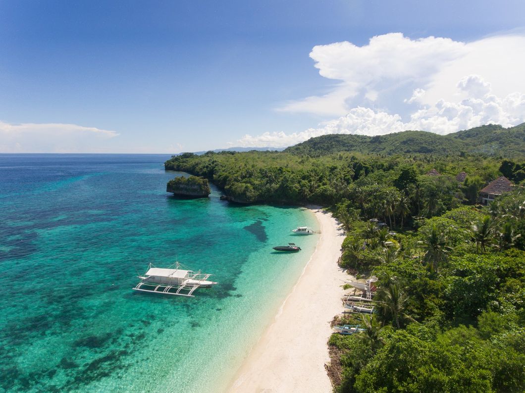 Tropical beach with turquoise water, white sand, lush green cliffs, and small boats offshore