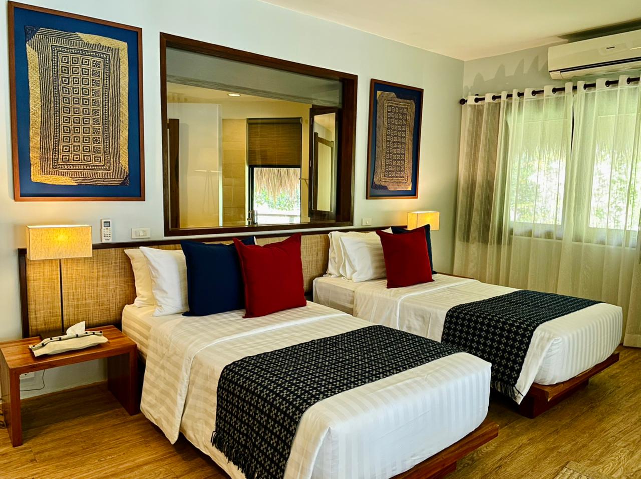 Bright bedroom with two twin beds, red and blue pillows, a large mirror, and soft curtains by the window