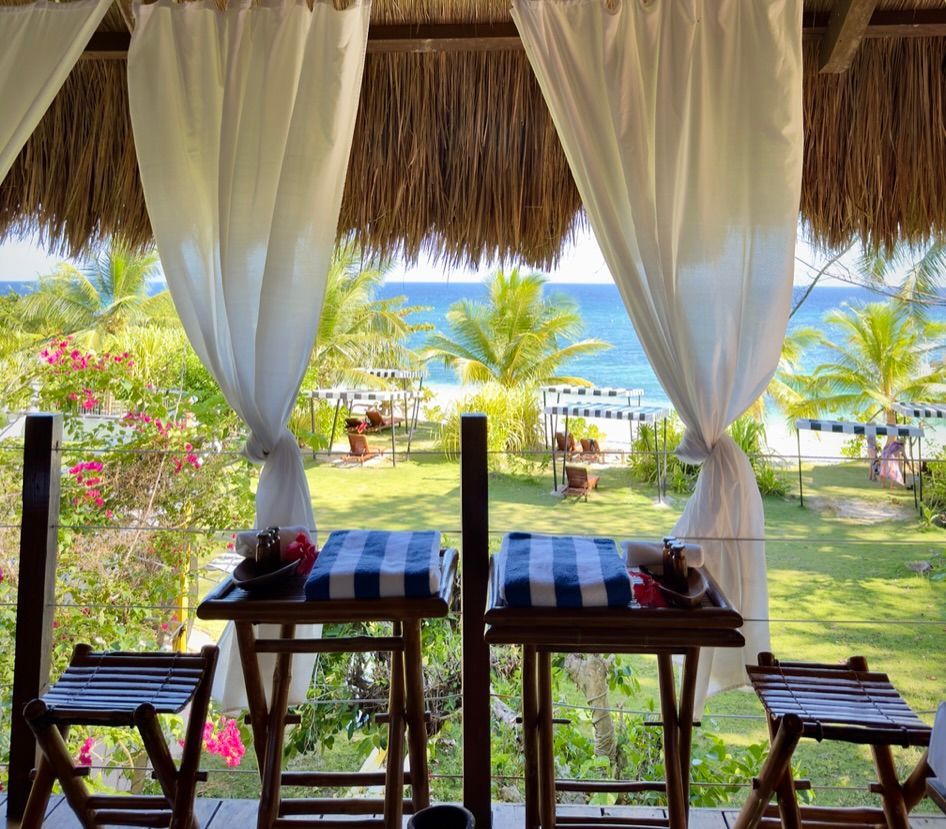 Beachfront cabana with white curtains, two small tables, and chairs overlooking palm trees and the ocean.