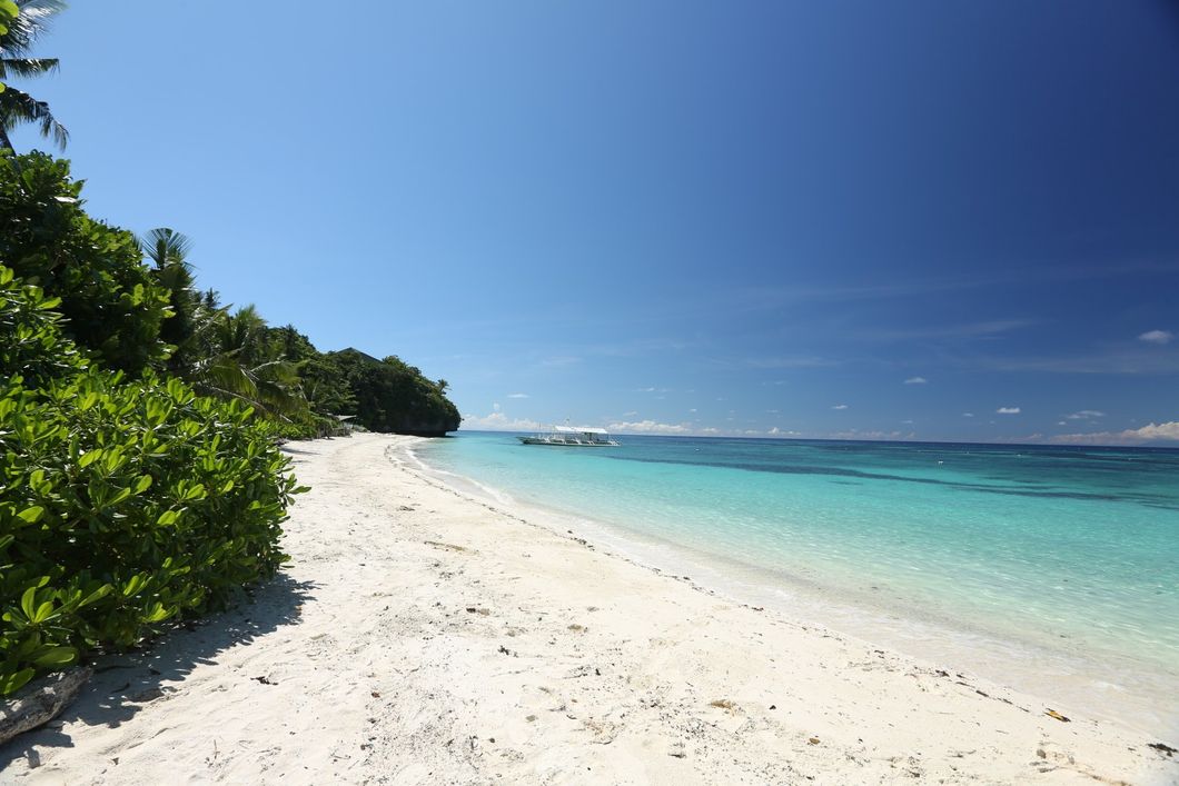 Sunny tropical beach with white sand, turquoise water, and lush green trees under a clear blue sky
