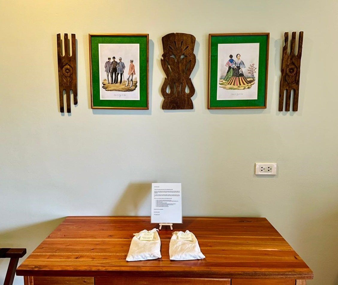 Wooden table with folded cloths and sign beneath framed art and carved wall hangings on a pale wall