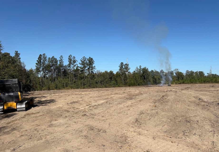 Cleared field with smoke rising, trees in background, small construction vehicle on the left.