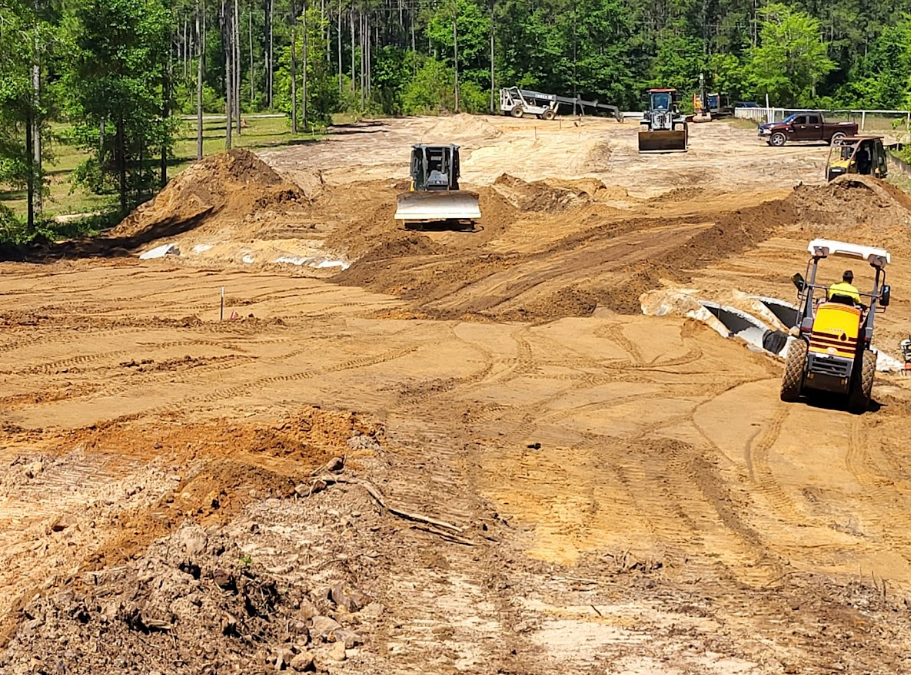 Construction site with heavy machinery grading the brown dirt, trees in the background.