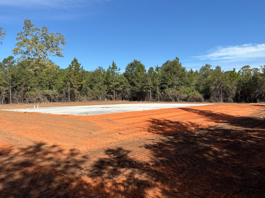 Cleared land with red soil in the foreground and a cleared, light-colored pad with trees and blue sky.