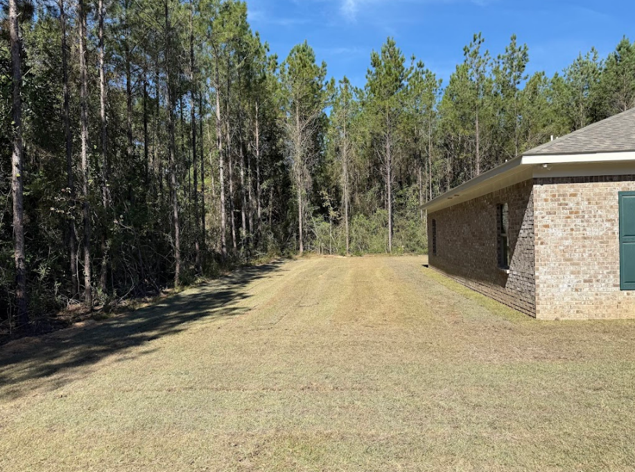 Side yard of a brick house with a treeline in the background and mowed grass in the foreground.