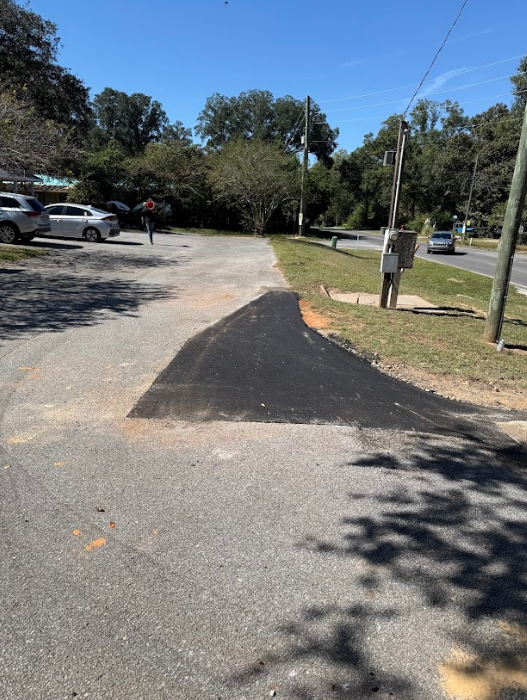 Newly paved asphalt section on a road, merging with the older surface. Trees and parked cars in the background.