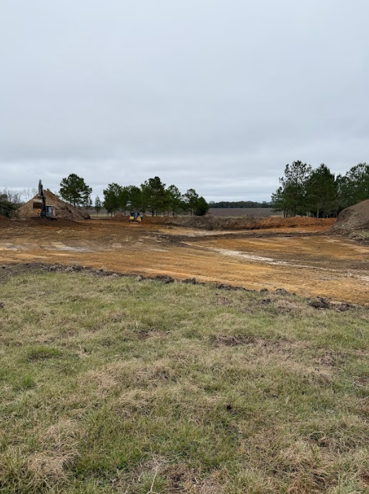 Construction site with dirt piles and machinery; grassy foreground and cloudy sky.