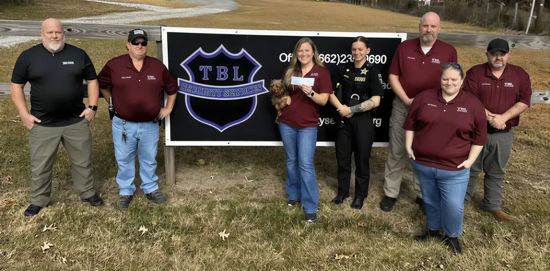 Seven people stand in a line before a sign with a police shield logo, holding a check in a grassy outdoor area.