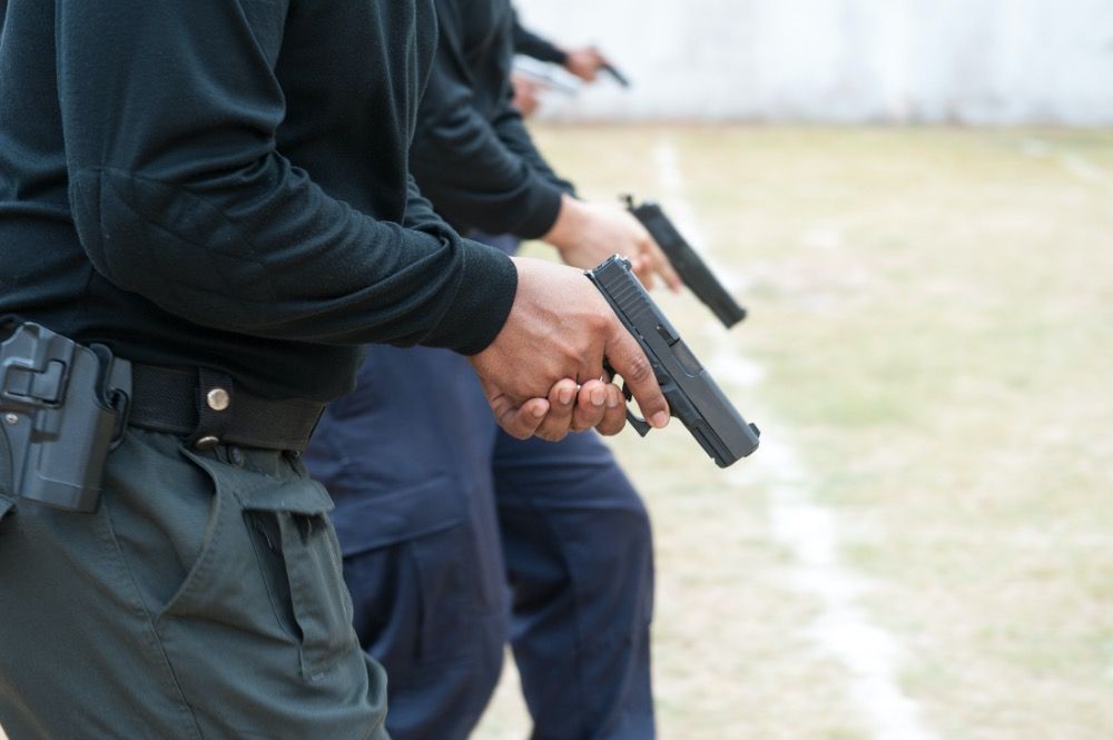 A line of individuals in dark uniforms standing on an outdoor range, holding handguns in a ready firing position.