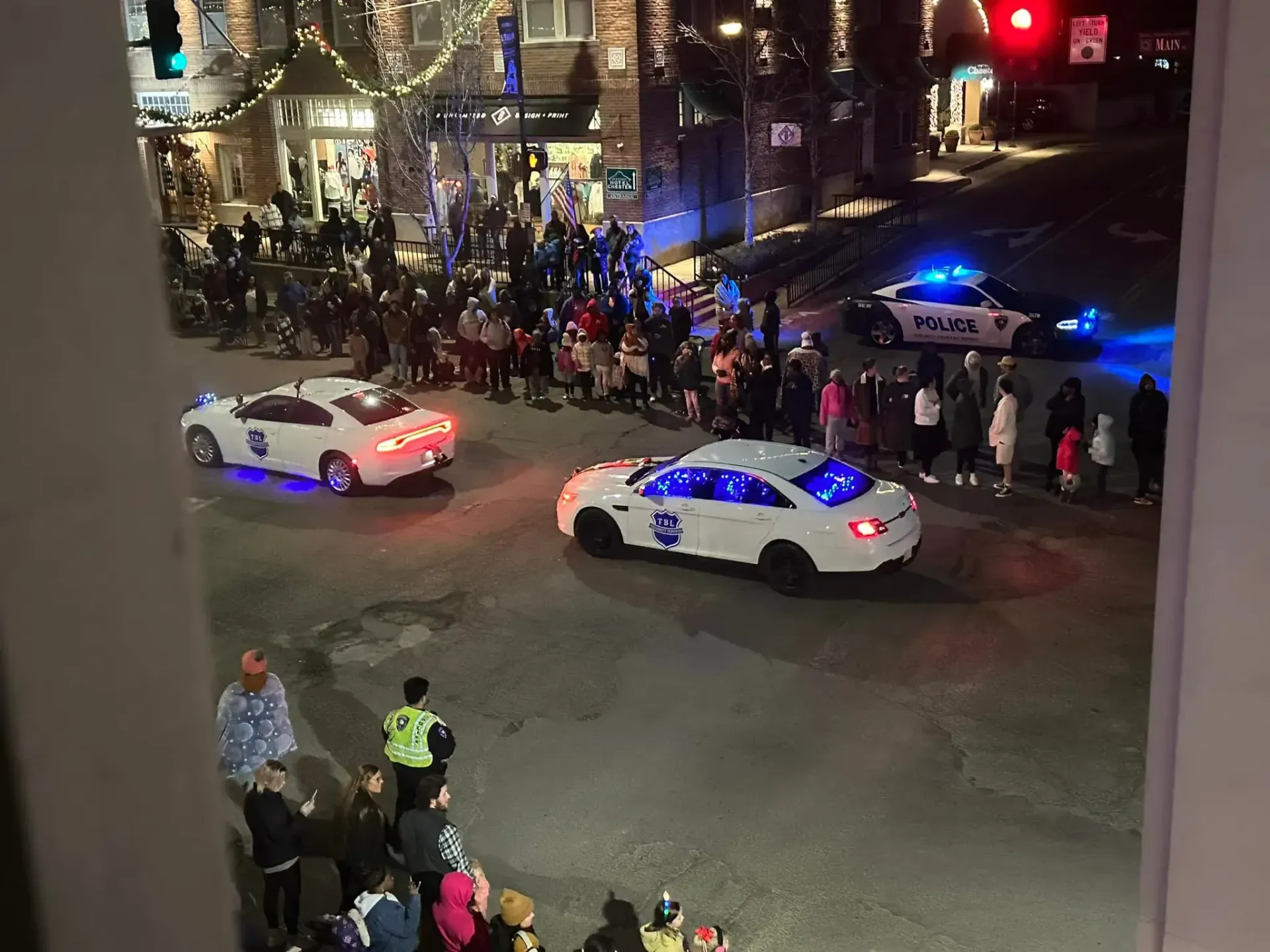 Police vehicles with flashing lights travel down a city street at night, passing a crowd gathered near storefronts.