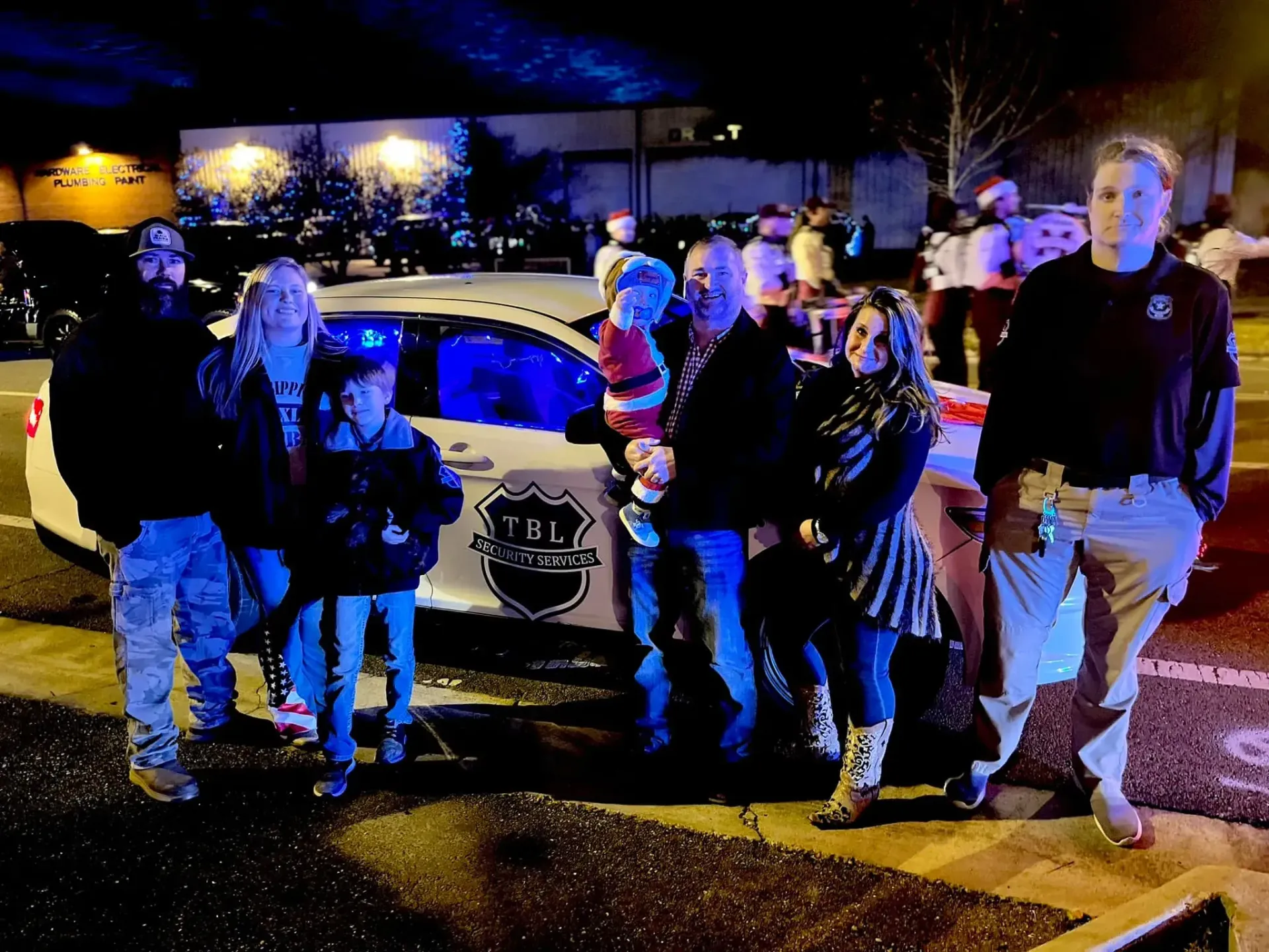 A group of people poses for a photo at night in front of a police cruiser with activated blue emergency lights.