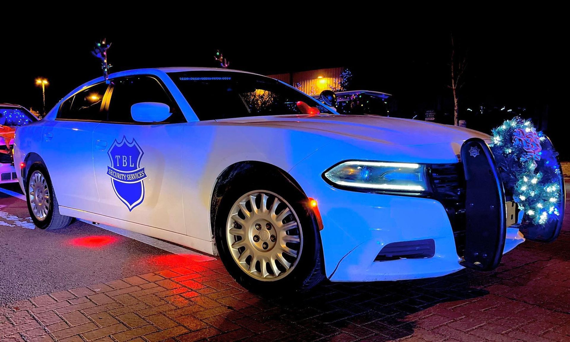 A white Dodge Charger police car decorated with festive Christmas lights and a badge emblem at night.