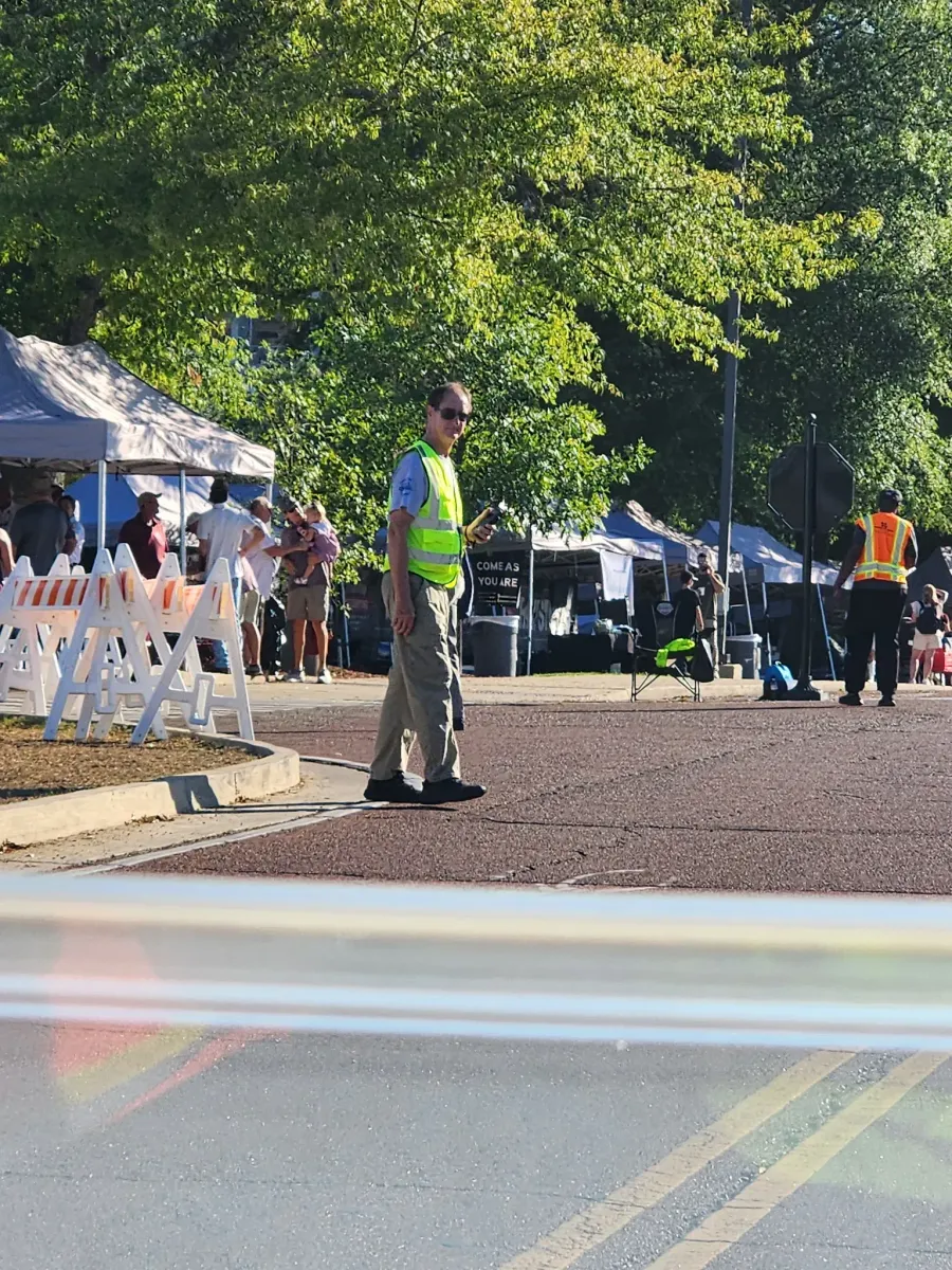 A uniformed crossing guard in a high-visibility vest stands on a dirt path near white barricades and event tents.