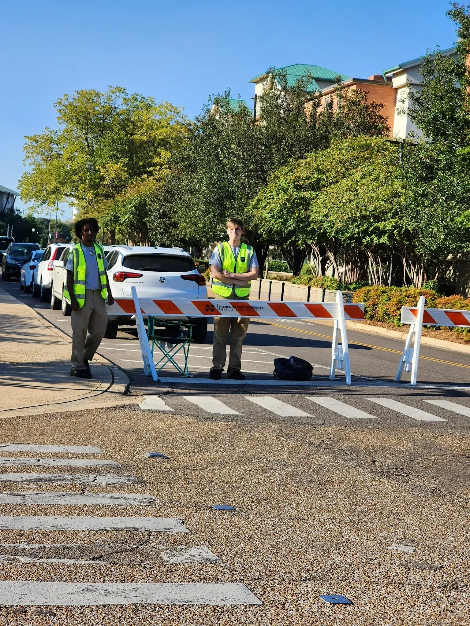 Two people in reflective safety vests stand behind a road barricade blocking a street in a tree-lined residential area.