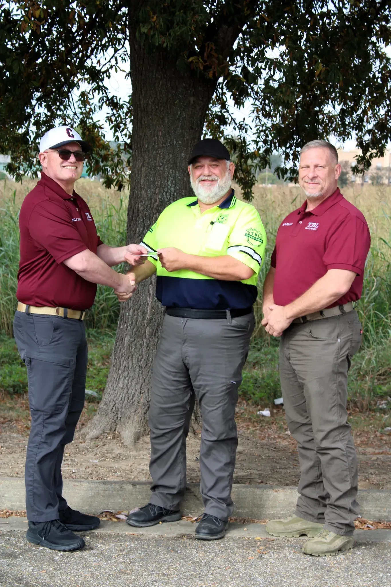 Three individuals in uniforms stand outdoors by a tree, with the person on the left handing a check to the person center.
