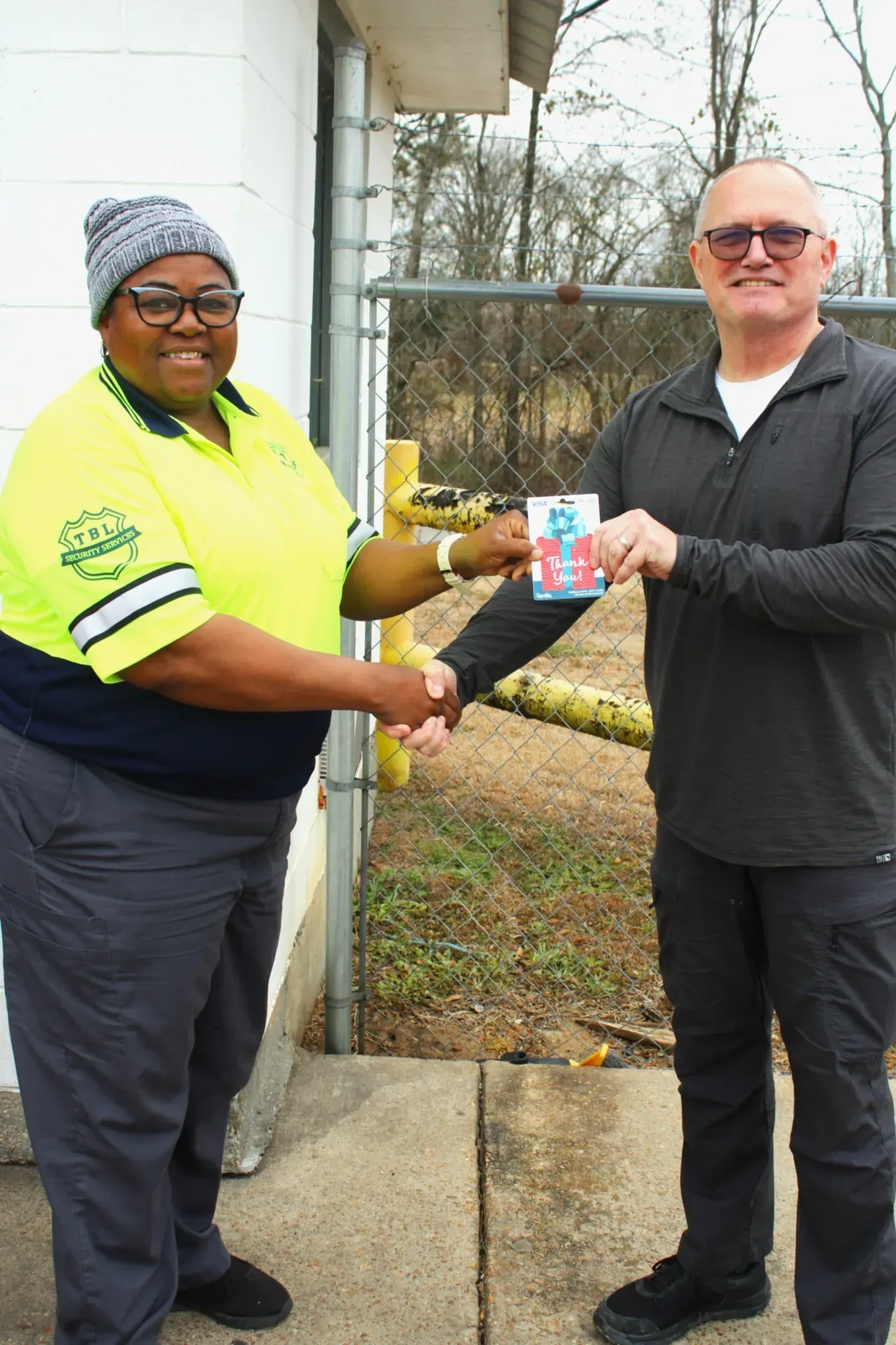 A person in a high-visibility yellow shirt shakes hands with someone in a black pullover while exchanging a small item.