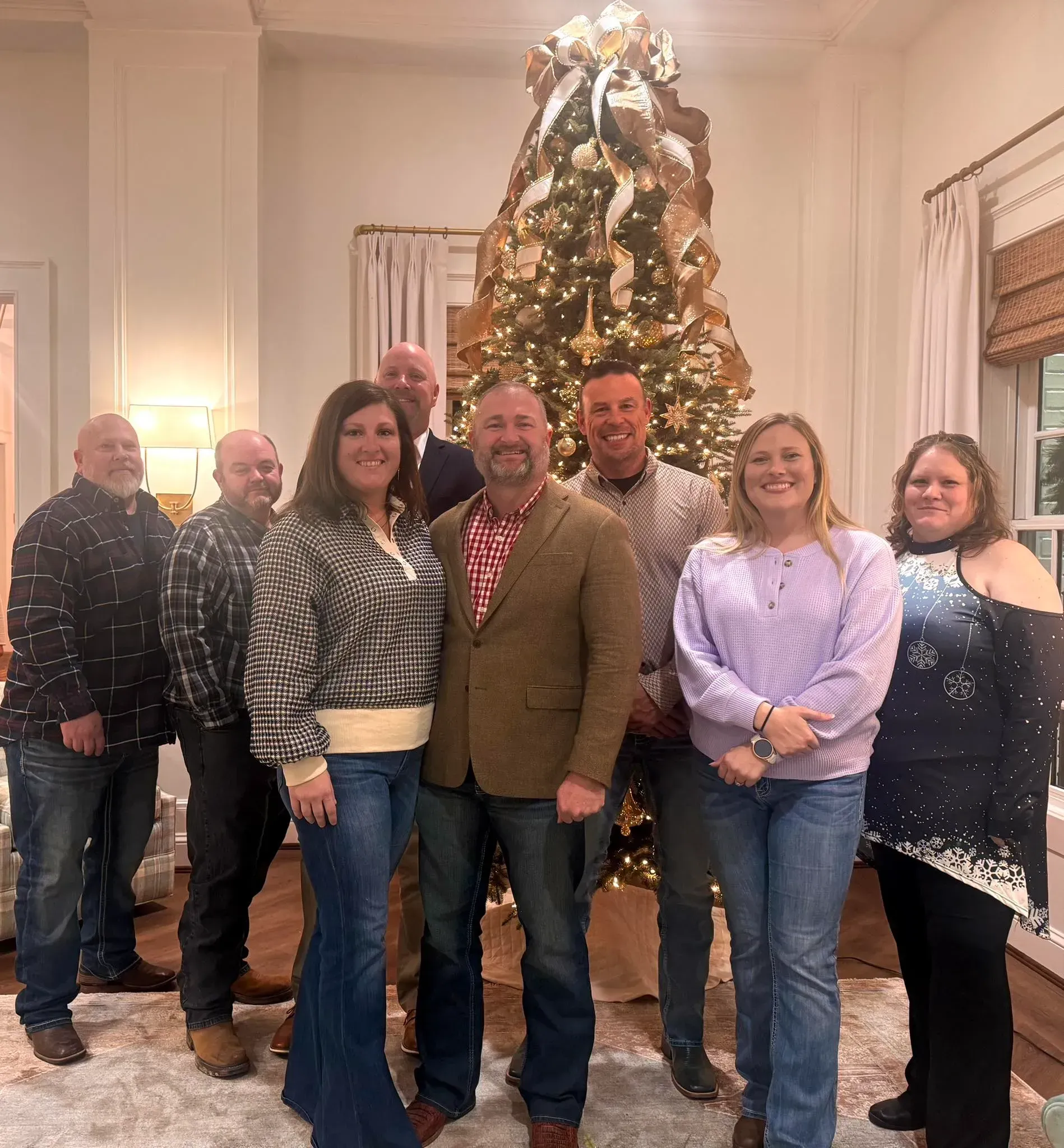 A group of eight people stand smiling in a living room in front of a large, decorated Christmas tree.