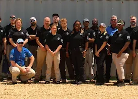 A group of staff members in matching uniforms pose together in a line outdoors against a corrugated metal wall.
