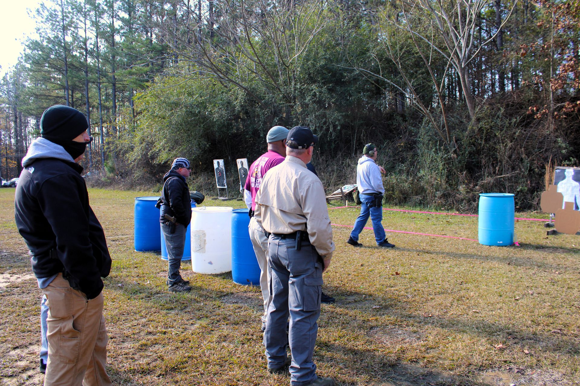 A group of people standing on an outdoor firing range near blue and white barrels and target silhouettes.