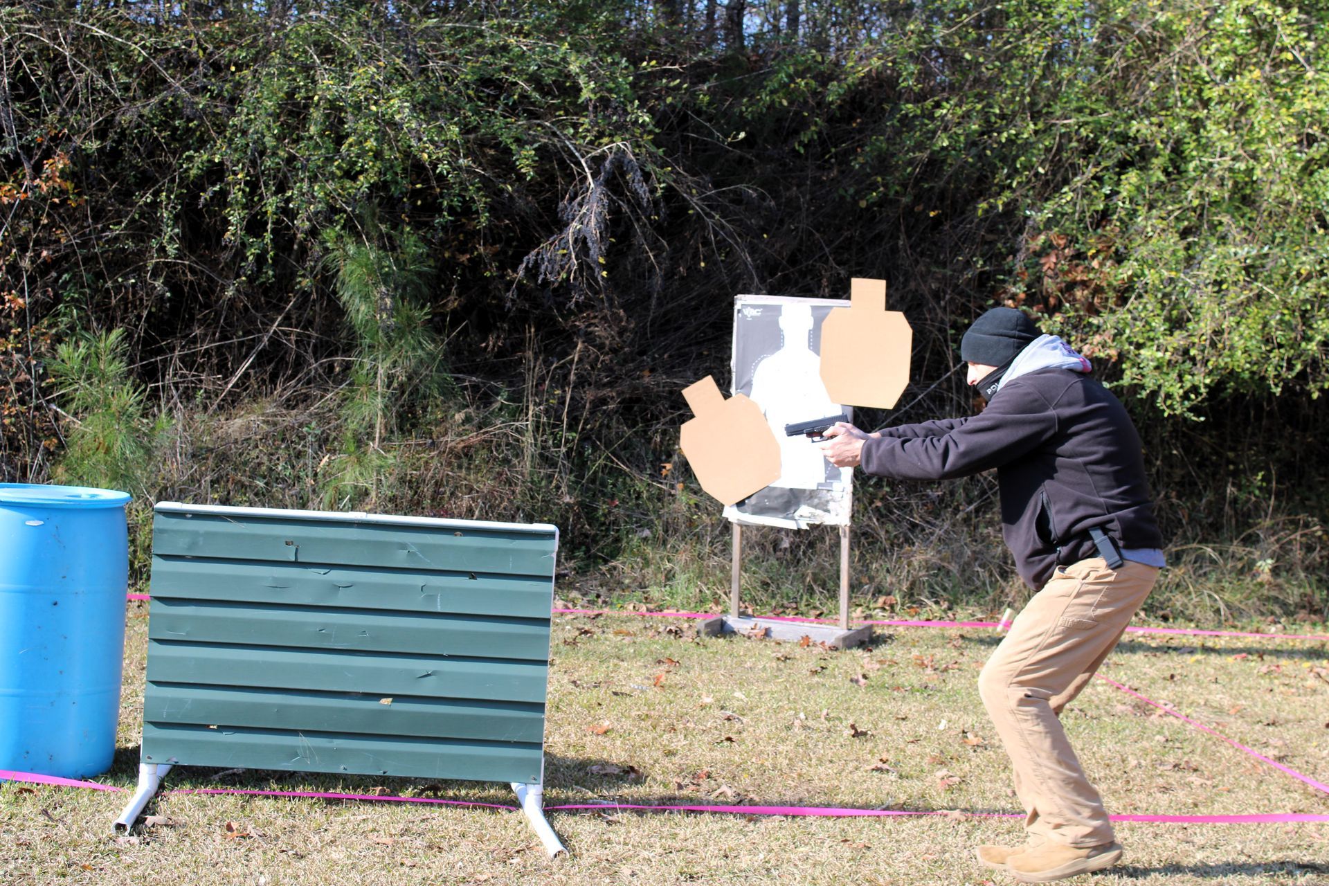 A person in a black jacket and tan pants shoots a handgun at paper targets on a stand in an outdoor range.