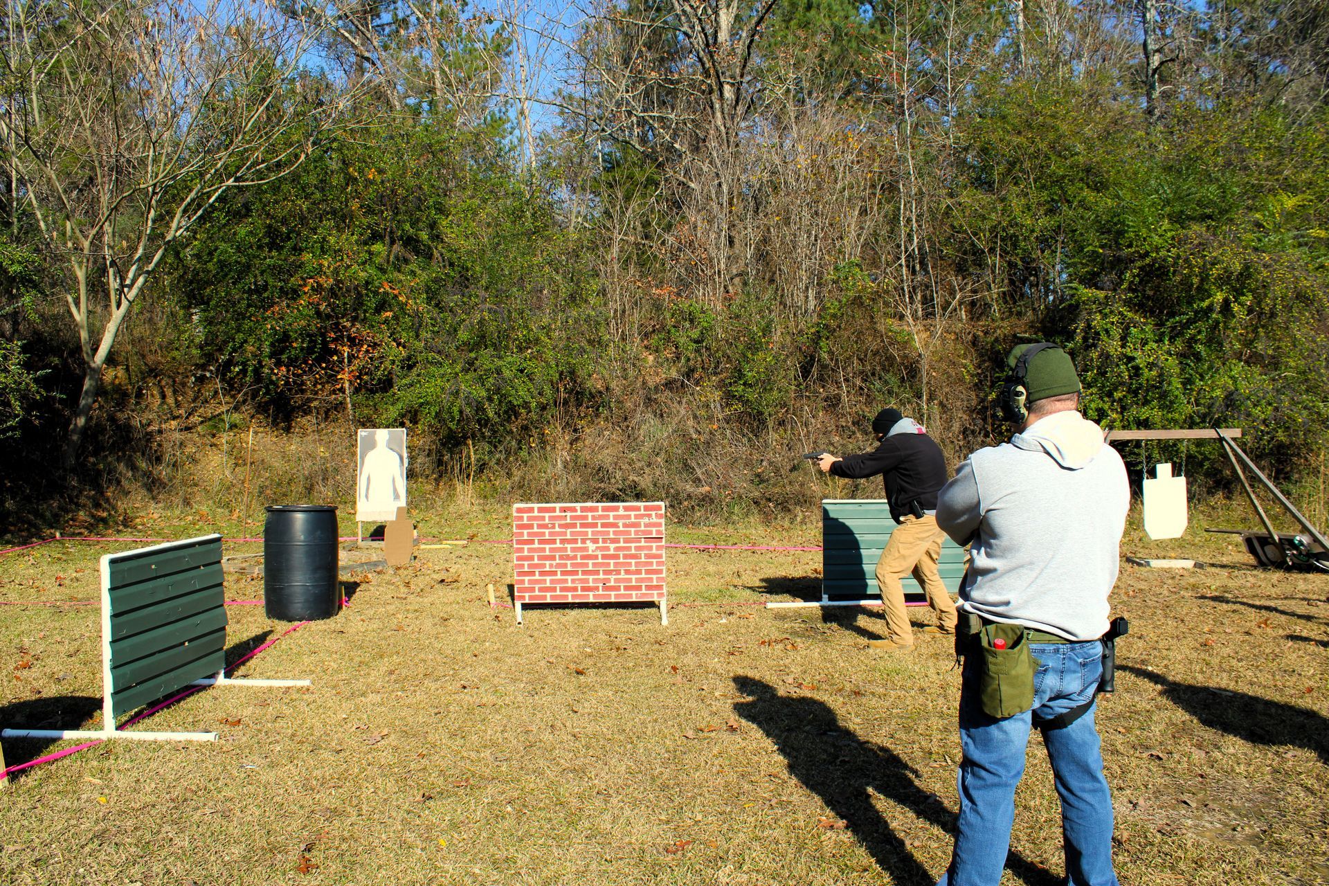 A person in a gray hoodie and blue jeans fires a handgun at targets on an outdoor shooting range.