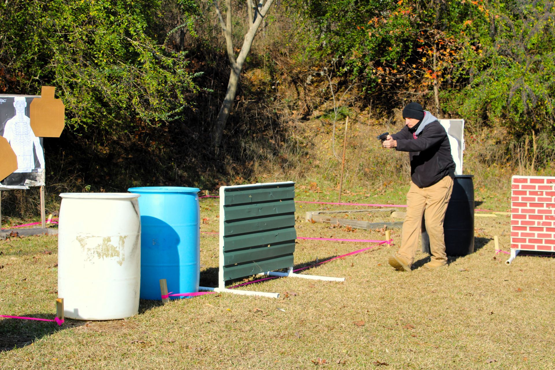 A person practicing at an outdoor shooting range, holding a handgun and standing near barrels and a target barrier.