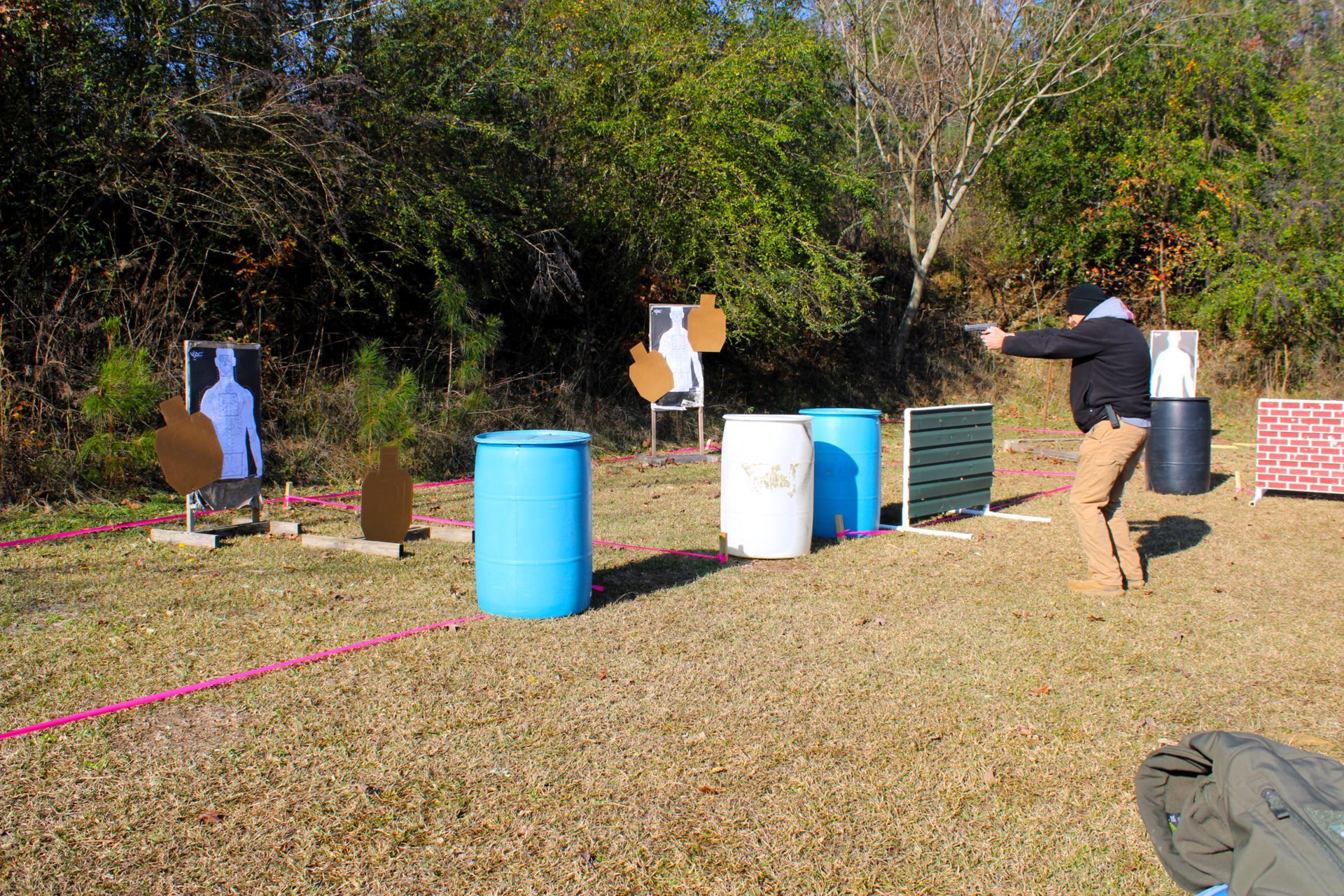 A person in tactical gear practices shooting at various target stands and barrels on an outdoor range.